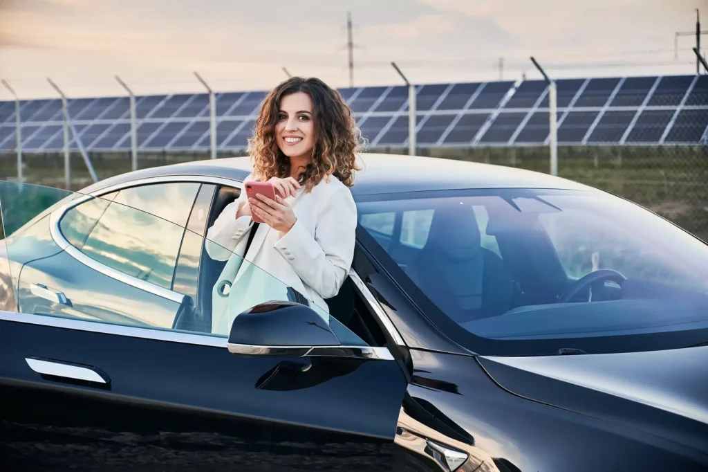 Cheerful woman standing by car and using smartphone.